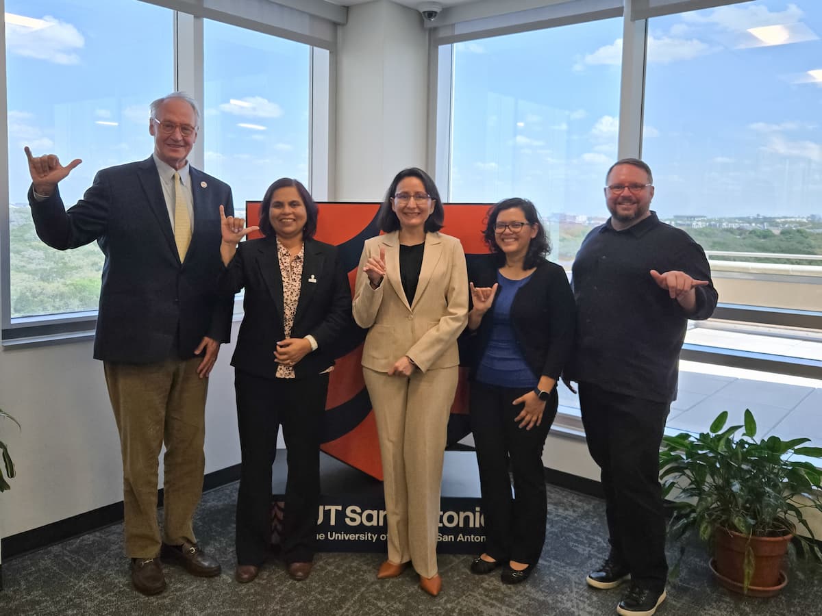 President Eighmy making a "birds up" gesture with HSRU postdoctoral fellows in front of UT San Antonio photo backdrop